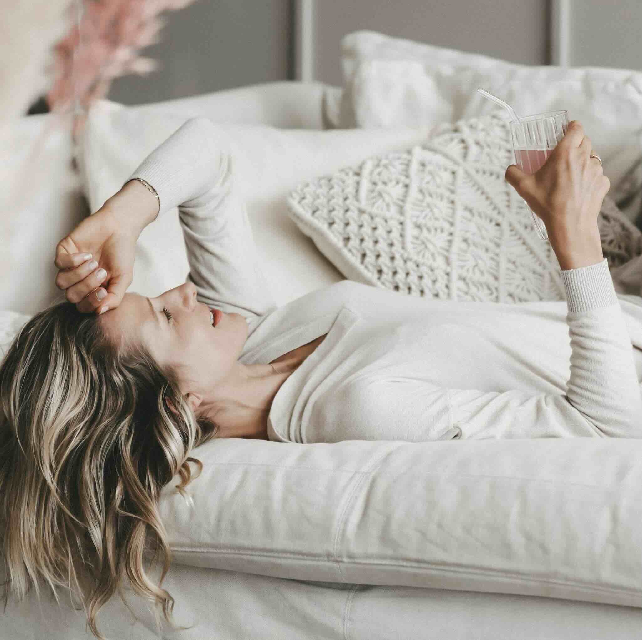 Frau entspannt sich in heller, gemütlicher Umgebung auf einem Sofa, trägt bequeme Kleidung und hält ein Glas. Die Szene strahlt Ruhe, Wellness und Selbstfürsorge aus.