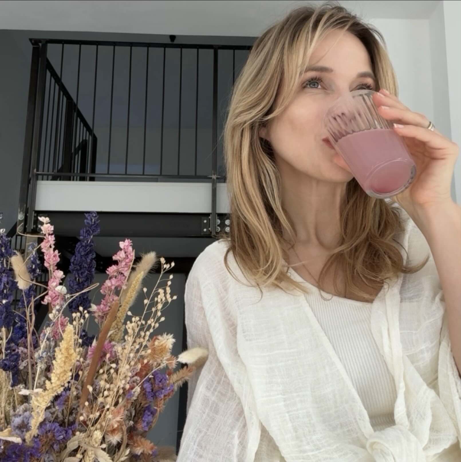 Frau mit blonden Haaren, Katja Will,  trinkt ein Glas Wildberry Beautyhacker Collagen-Drink, während sie entspannt in einem hellen Raum sitzt. Im Vordergrund steht ein dekorativer Strauß aus Trockenblumen in warmen Naturtönen.